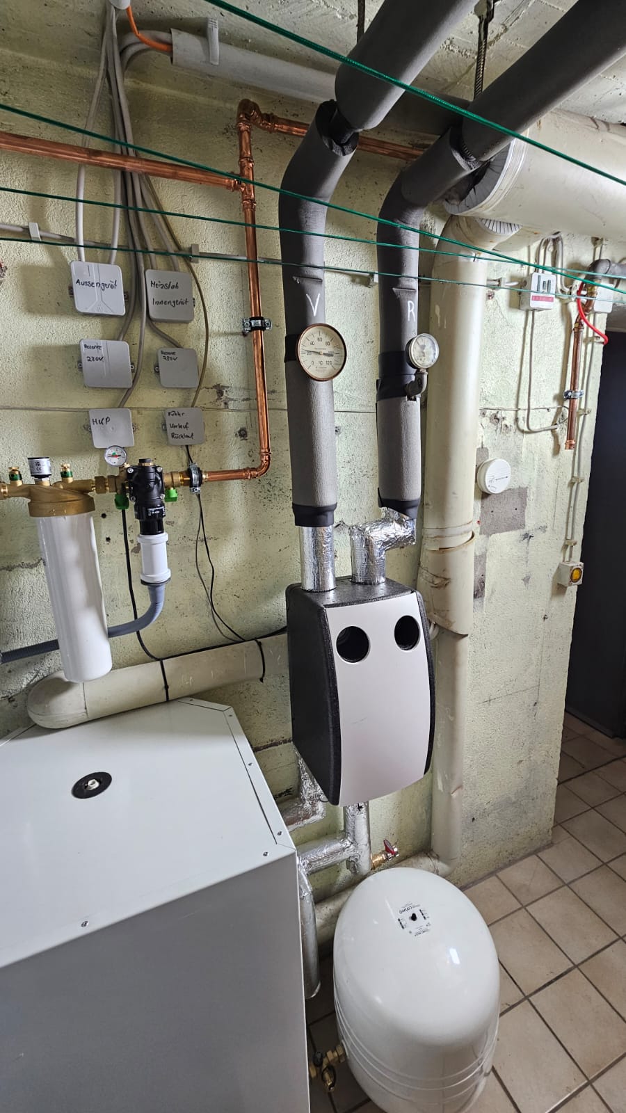 Wärmepumpen-Inneneinheit mit Verrohrung, Druckanzeige und Filtertechnik in einem gefliesten Heizungsraum mit Beschriftungen.